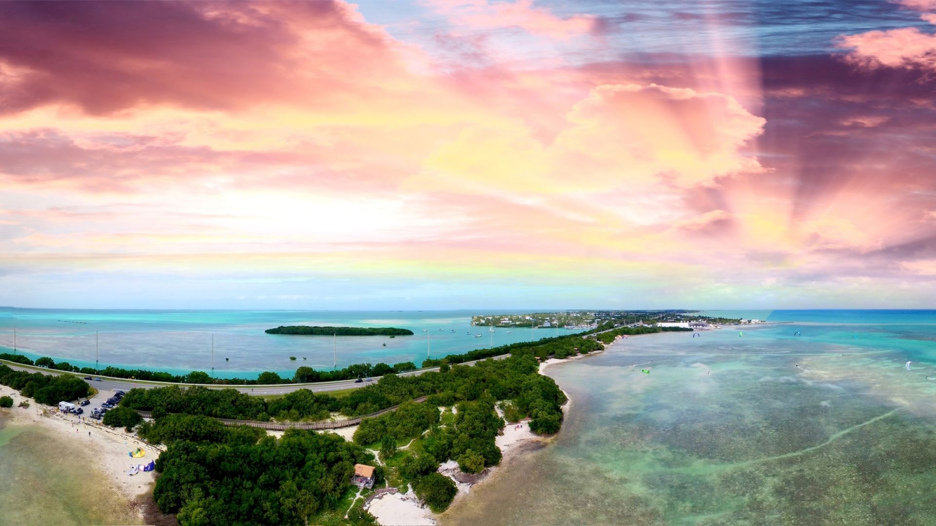 Aerial view of a tropical beach and lagoon with boats, greenery, and a colorful sunset sky overhead.