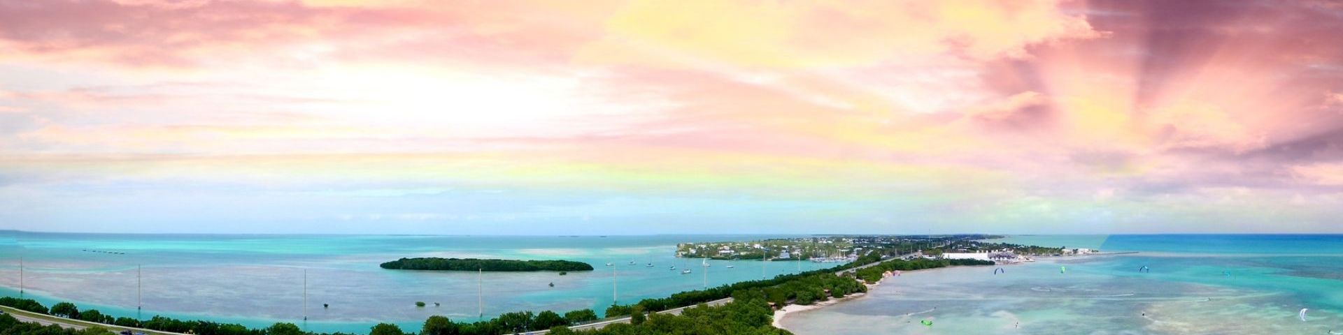 Aerial view of a tropical beach and lagoon with boats, greenery, and a colorful sunset sky overhead.