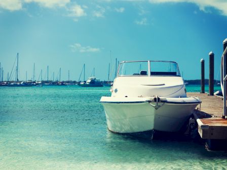 A white boat is docked at a pier with a clear blue sky and multiple sailboats in the background on a calm, turquoise sea.