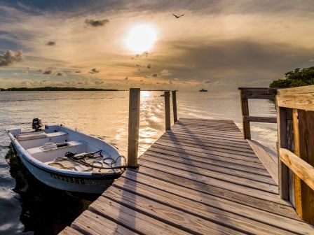 A small boat is moored beside a wooden dock at sunset, with calm water and a distant shore under a cloudy sky. A bird is flying overhead.
