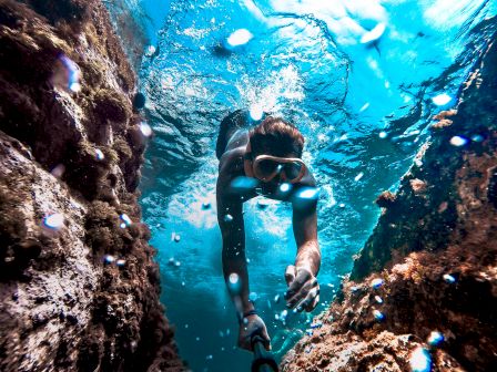 A diver swims through a narrow underwater passage between rocks, surrounded by clear blue water and bubbles, wearing goggles and a snorkel.