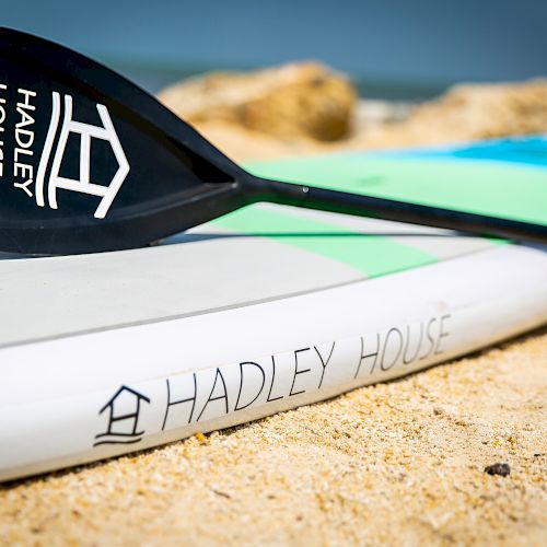 A paddle and a surfboard with "Hadley House" branding lay on a sandy beach with some rocks in the background.