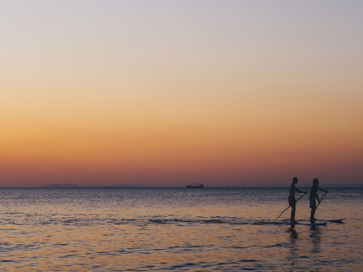 Two people are paddleboarding on calm water during a vibrant sunset, with the horizon and a distant ship visible in the background.
