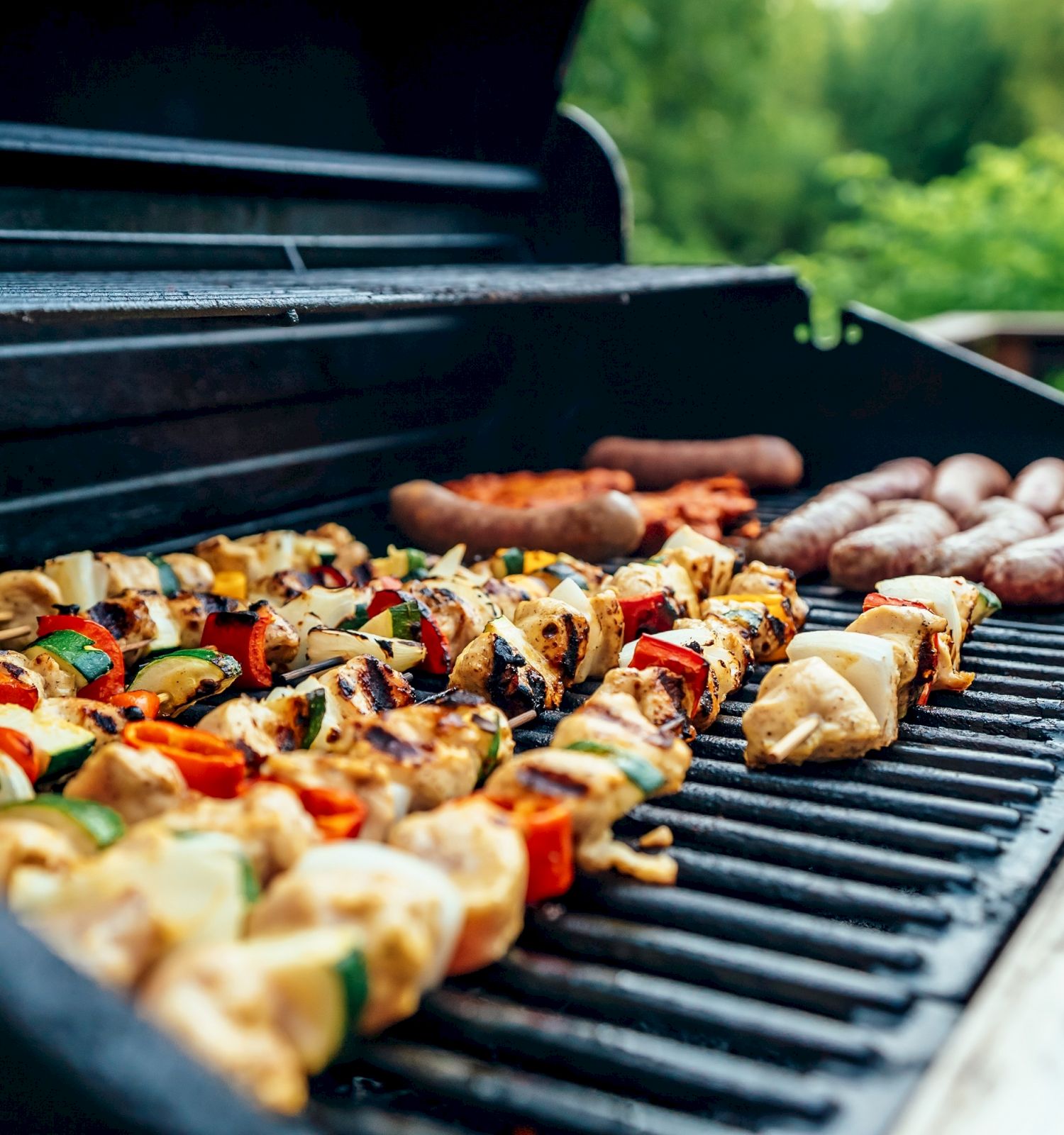 A grill with skewered vegetables and sausages being cooked, with a green, natural background visible and a deck railing in the back.