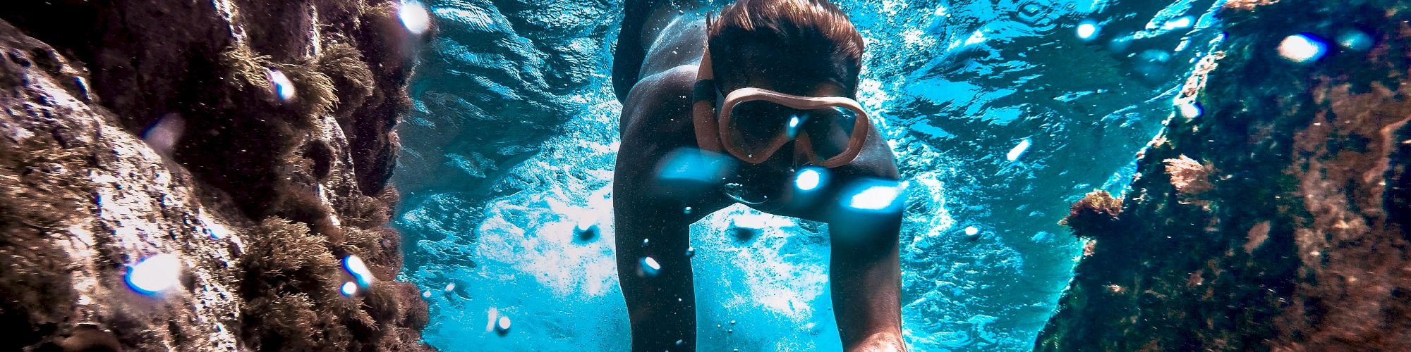 A person in snorkeling gear is underwater, swimming through a rocky underwater channel with clear blue water surrounding them.