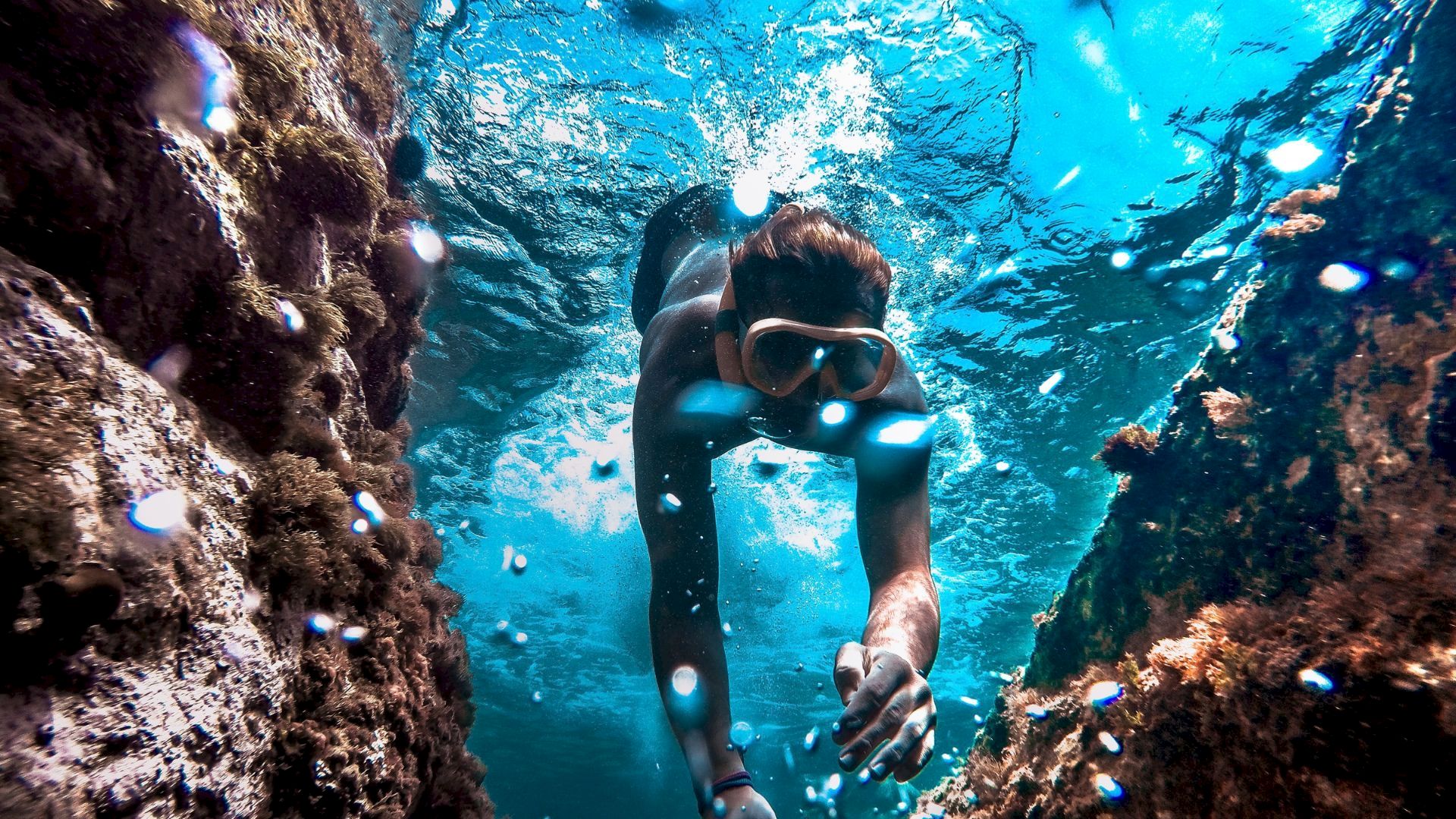 A scuba diver swims through clear blue water near a coral reef, wearing a mask and breathing underwater.