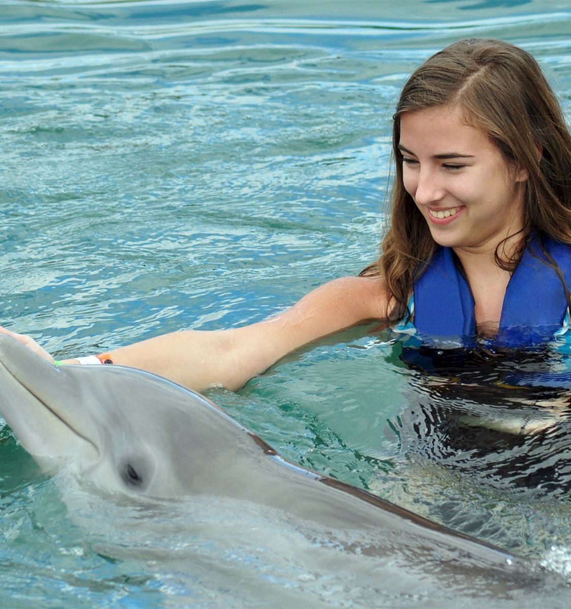 A woman in a blue vest floats in the ocean, smiling as she holds a dolphin by the snout.