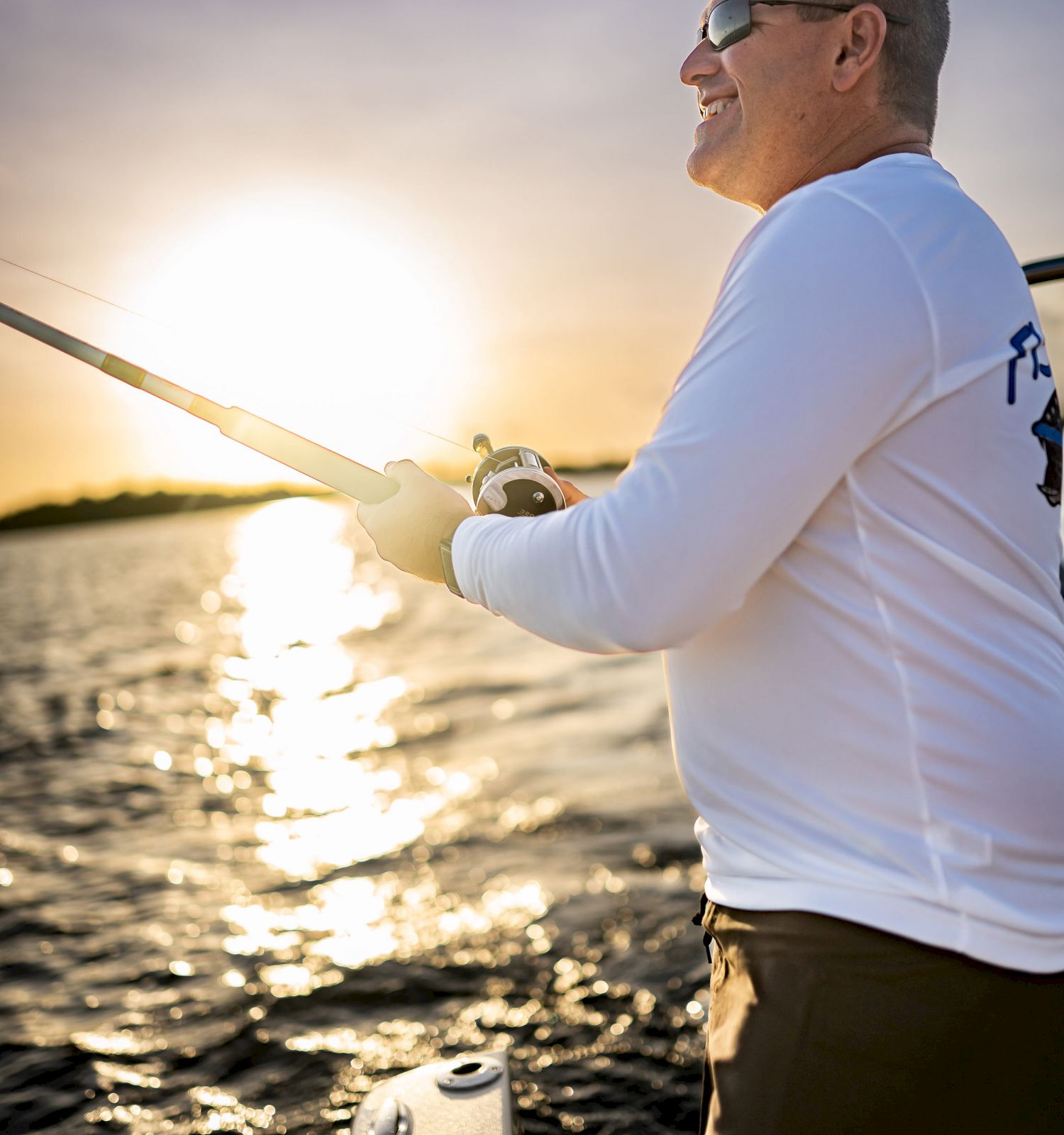 A person is fishing on a boat during sunset, wearing a long-sleeve shirt and sunglasses, with water and the sun in the background.