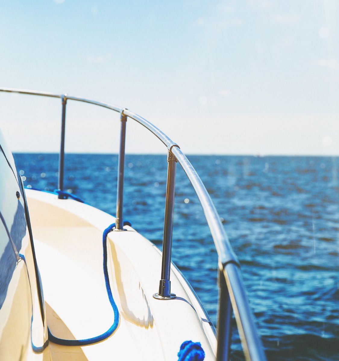 A boat&rsquo;s bow and rails stretch over open blue ocean, with sunlight and distant horizon under a clear sky.