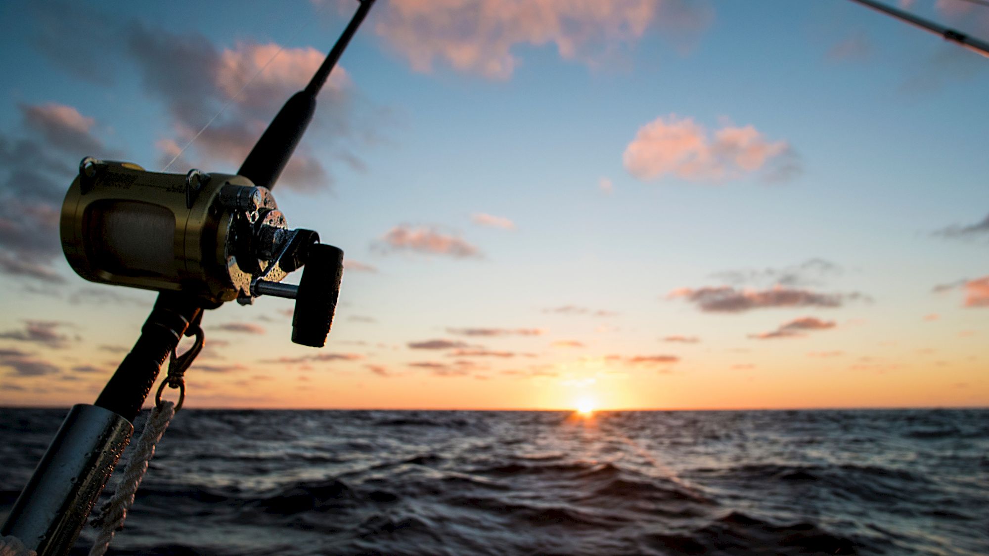 The image shows a fishing rod mounted on a boat with the sun setting over the ocean in the background, casting warm colors across the sky.