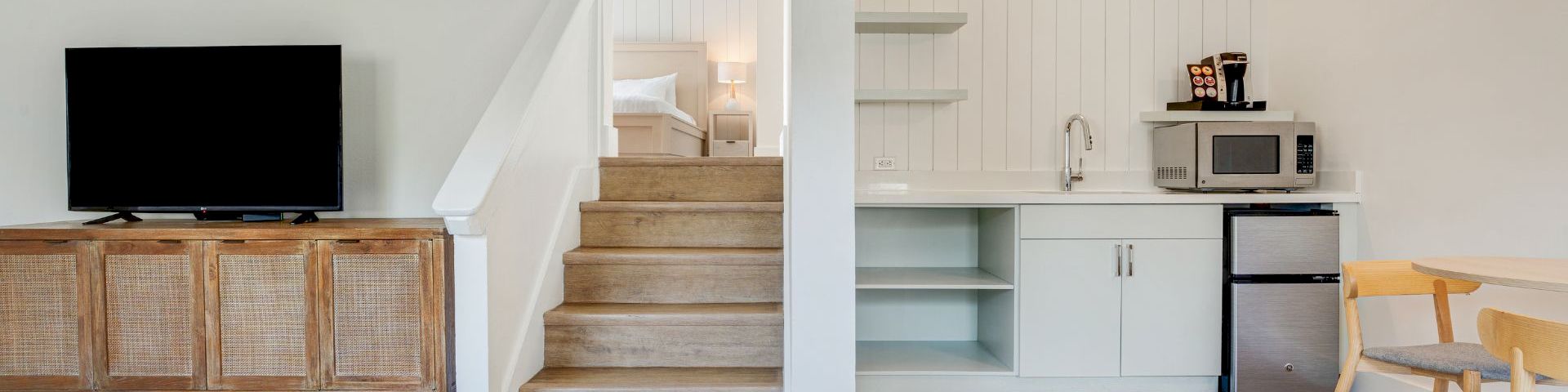 Bright modern living area with a TV, stairs to a bedroom, and a built-in kitchenette with microwave and coffee machine.