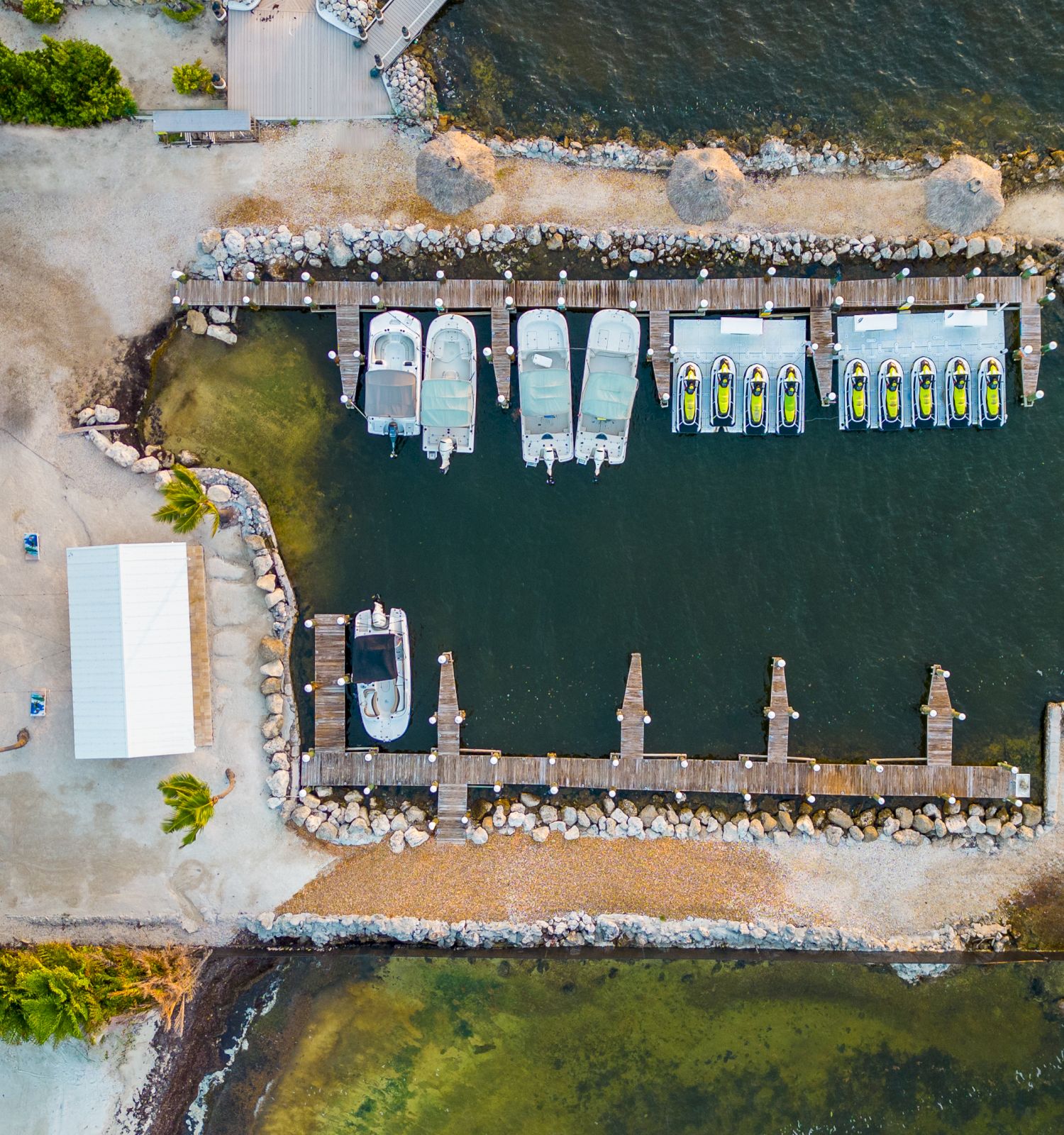 Aerial view of a marina with several boats docked, surrounded by rocks and water, adjacent to a white building and parking lot.