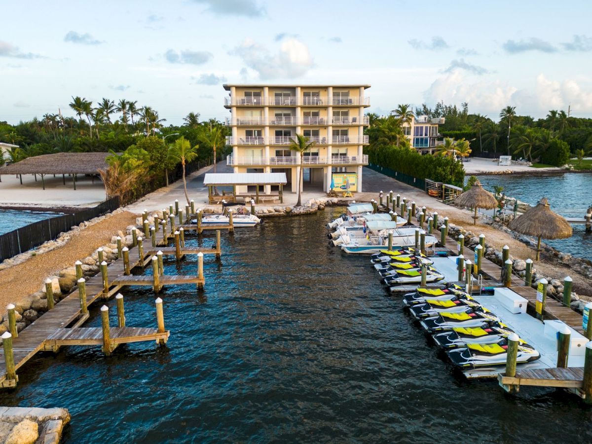 A tropical resort waterfront with a multi-story hotel, docks, and rows of bright jet skis by the bay, under clouds.