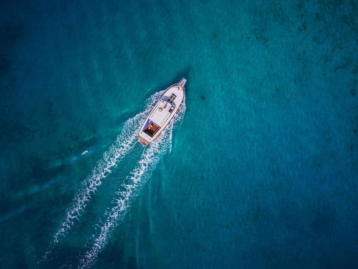 Aerial view of a small white motorboat cruising through clear turquoise water, leaving a foamy wake behind it.