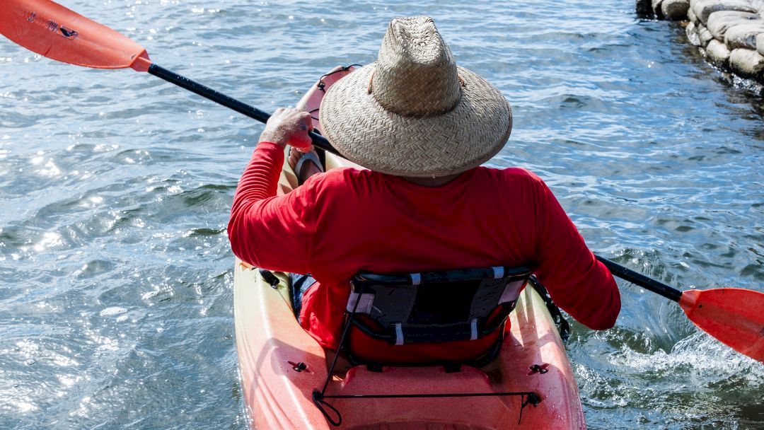 A kayaker in a straw hat paddles a red kayak on open water under a bright sky.