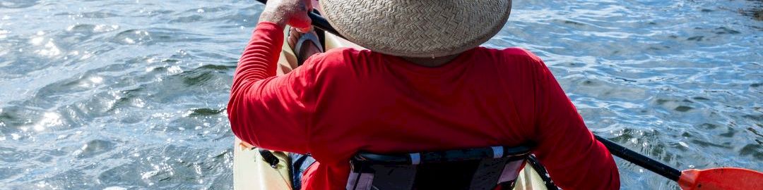 A kayaker in a straw hat paddles a red kayak on open water under a bright sky.
