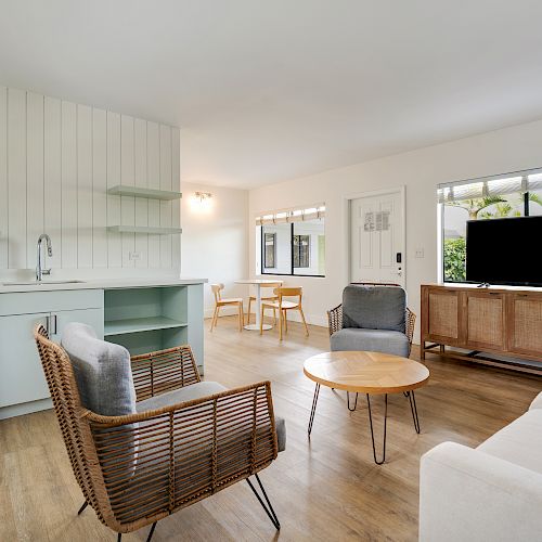 A modern living room with a kitchenette, seating area, TV on a wooden stand, a coffee table, and a small dining area with a table and chairs by the window.