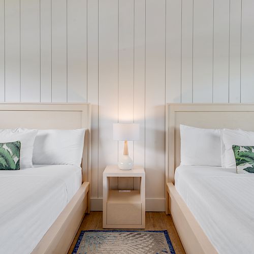 The image shows a bedroom with two white beds, each with green leaf-patterned pillows, a small nightstand in between, and a table lamp on it.