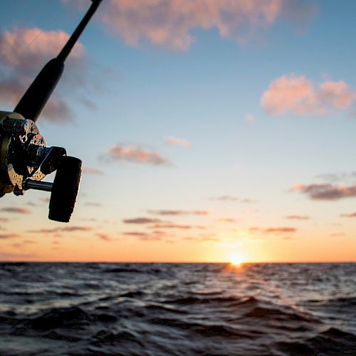 A fishing reel and rod are set against the backdrop of a calm ocean at sunset, with a few clouds scattered in the sky.