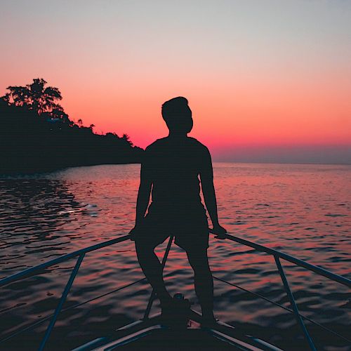 A silhouetted person stands at the bow of a boat looking towards the horizon during a vibrant sunset over calm waters and an island in the distance.