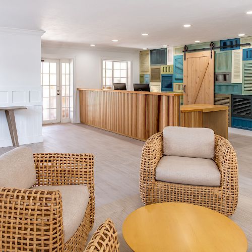 A modern lobby area featuring wicker chairs, a round wooden table, a reception desk, and a colorful wall design with shutter patterns, well-lit.