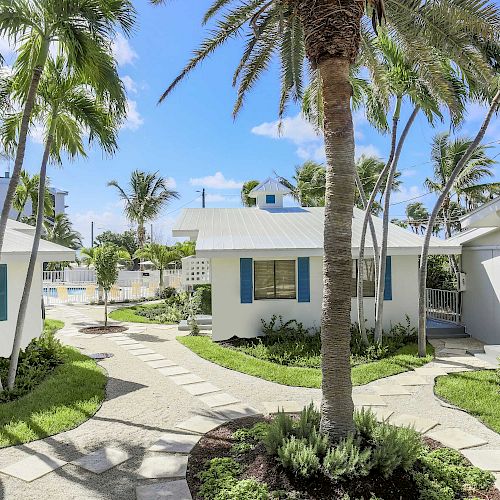 A courtyard with small, white cottages, surrounded by palm trees and a clear blue sky, featuring pathways leading to each cottage.