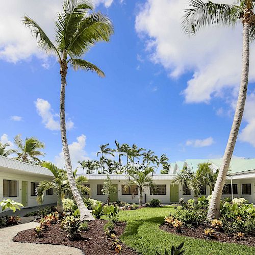 A courtyard with palm trees, white buildings, and a blue sky with clouds. There are walkways and tropical plants around the area.