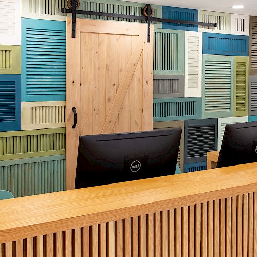 The image shows a reception desk with two computer monitors and a decorative wall featuring various colorful shutter panels and a wooden door.