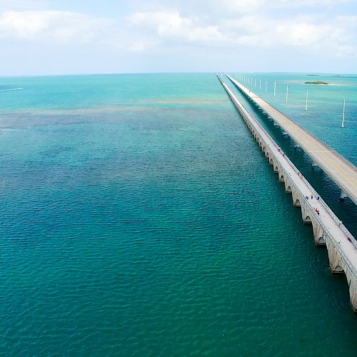 This image depicts a long bridge stretching over turquoise ocean water, connecting small islands under a partly cloudy sky, ending the sentence.