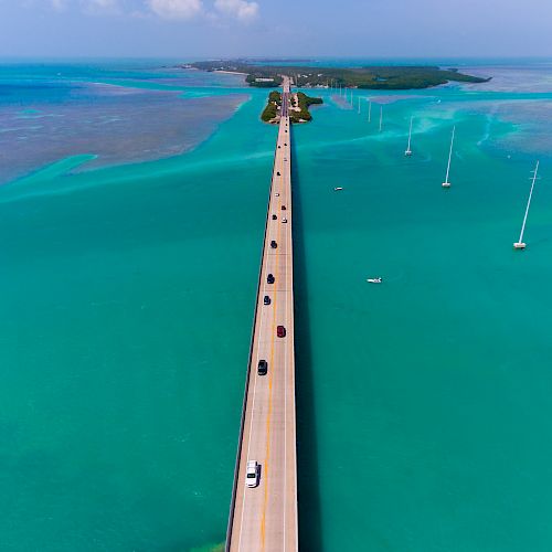 The image shows a long bridge over turquoise water with several cars traveling on it, leading to a distant island.