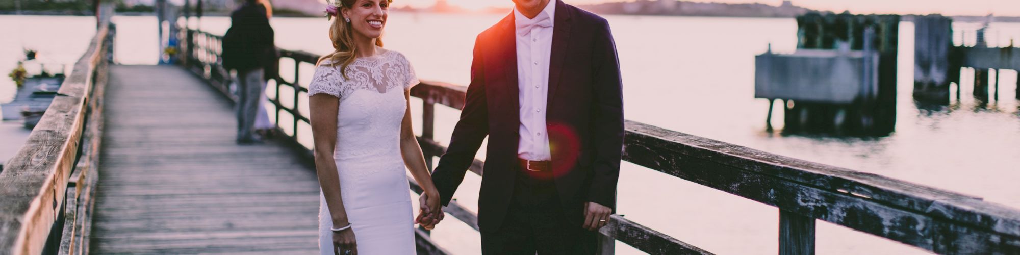 A couple stands on a pier at sunset, dressed in wedding attire, hand in hand, with water and a beautiful horizon in the background.