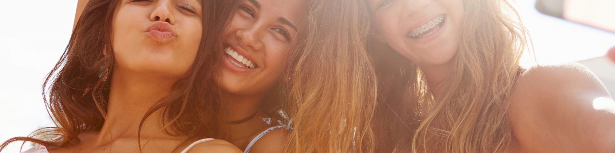 Three women pose outdoors, smiling and making playful gestures. The background is bright and sunlit.