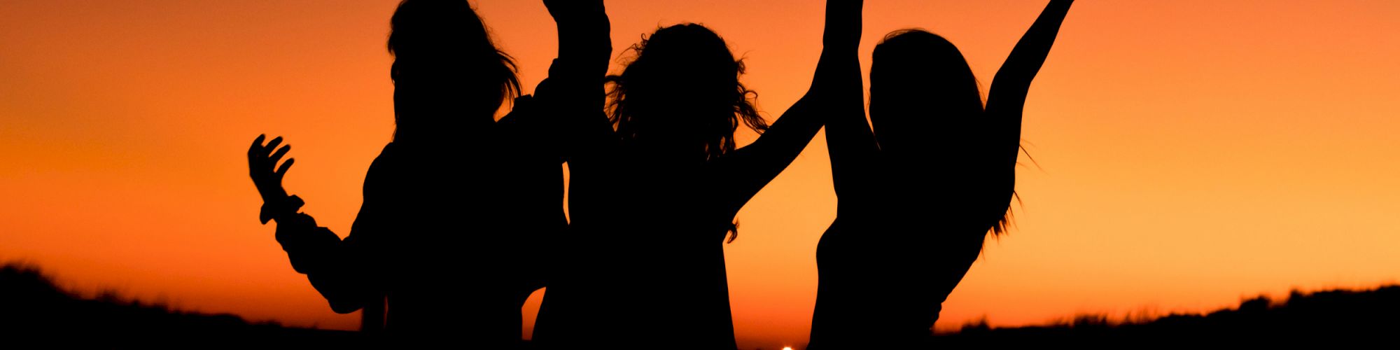 Three silhouetted individuals are dancing or celebrating with their hands raised against a vibrant sunset sky.