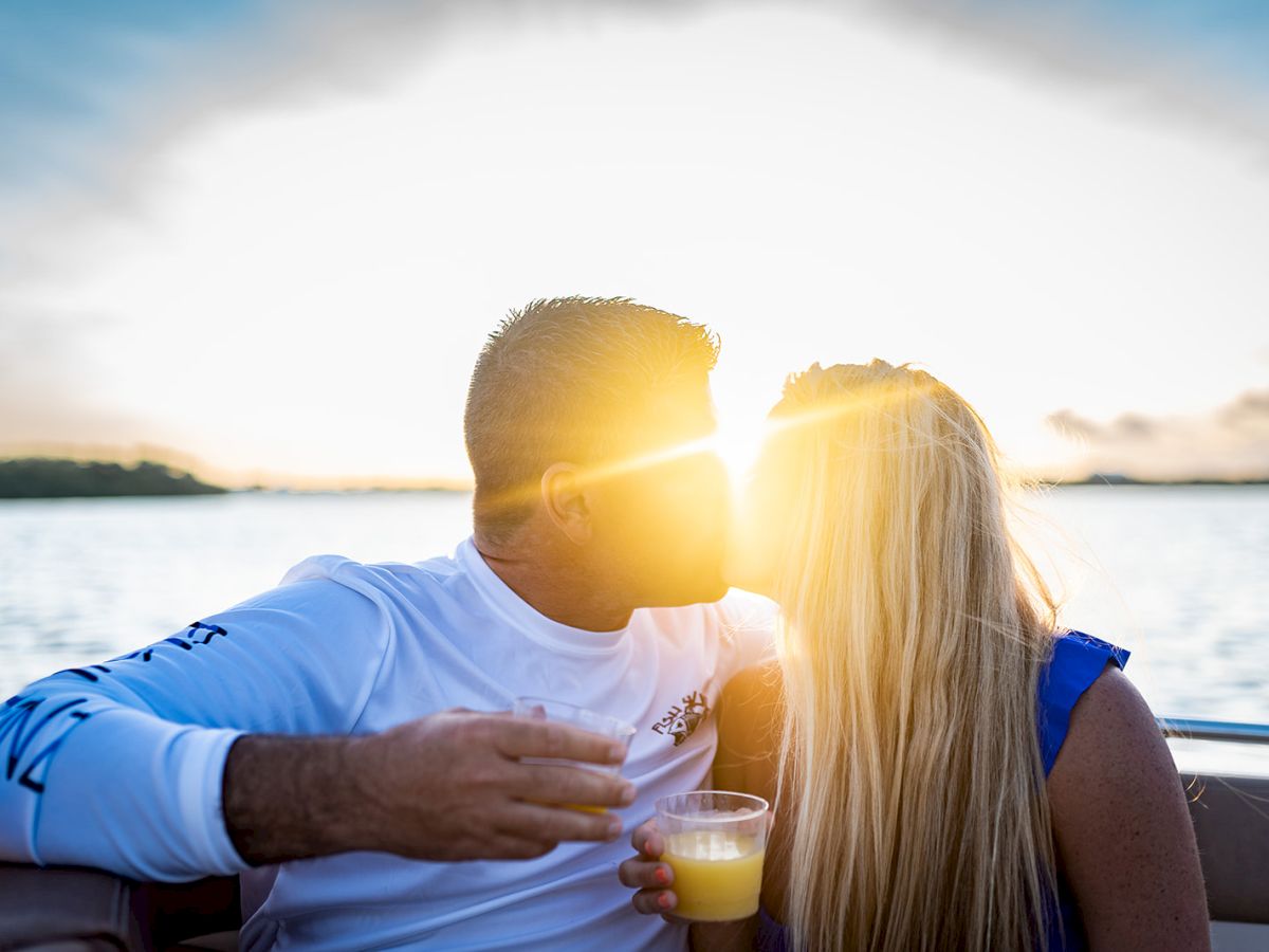 A man and blonde woman share a kiss on a boat at sunset by the water, holding drinks and smiling together.
