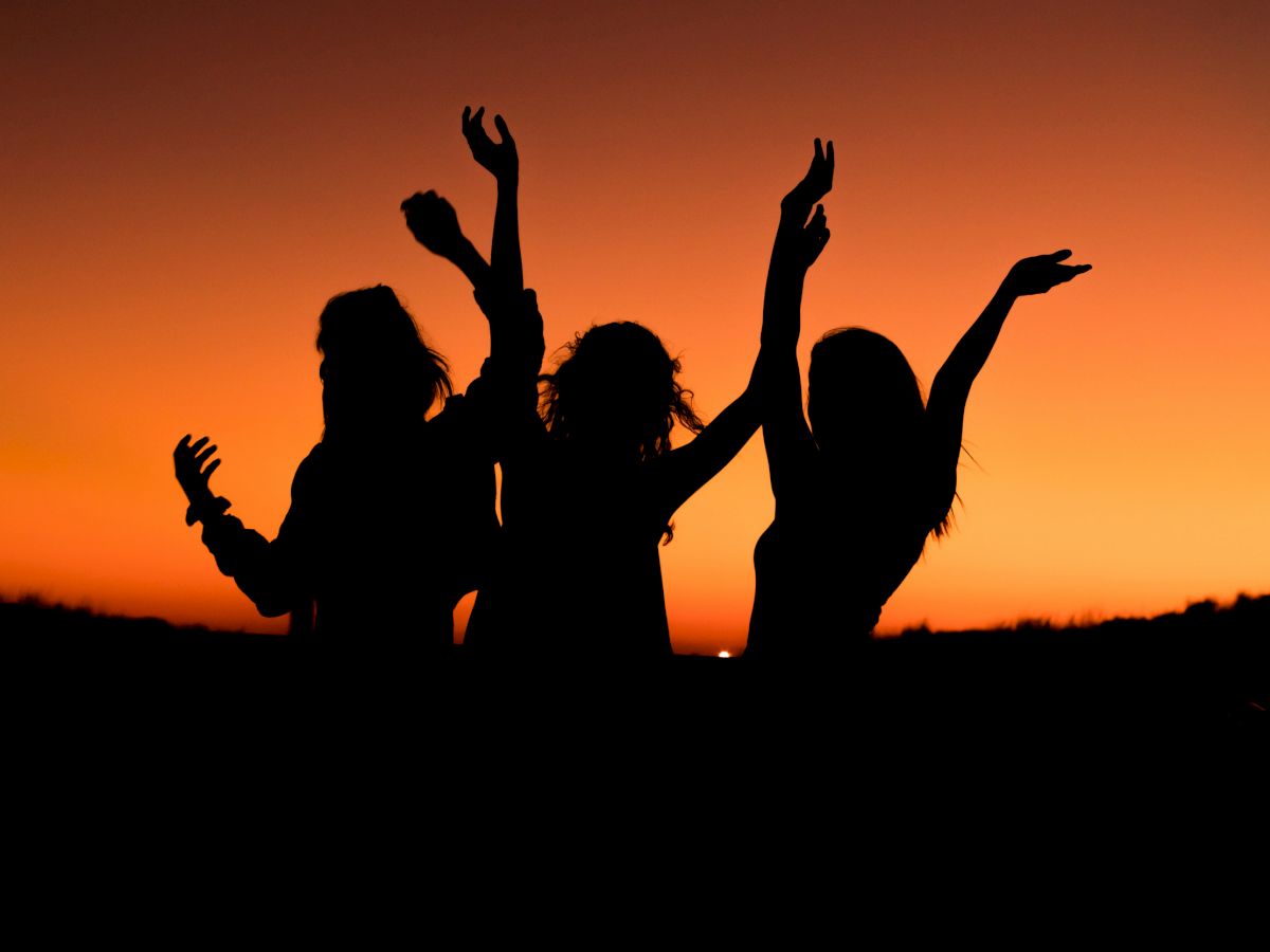 Three silhouetted individuals are dancing joyfully against a vivid orange sunset sky, their arms raised in celebration.