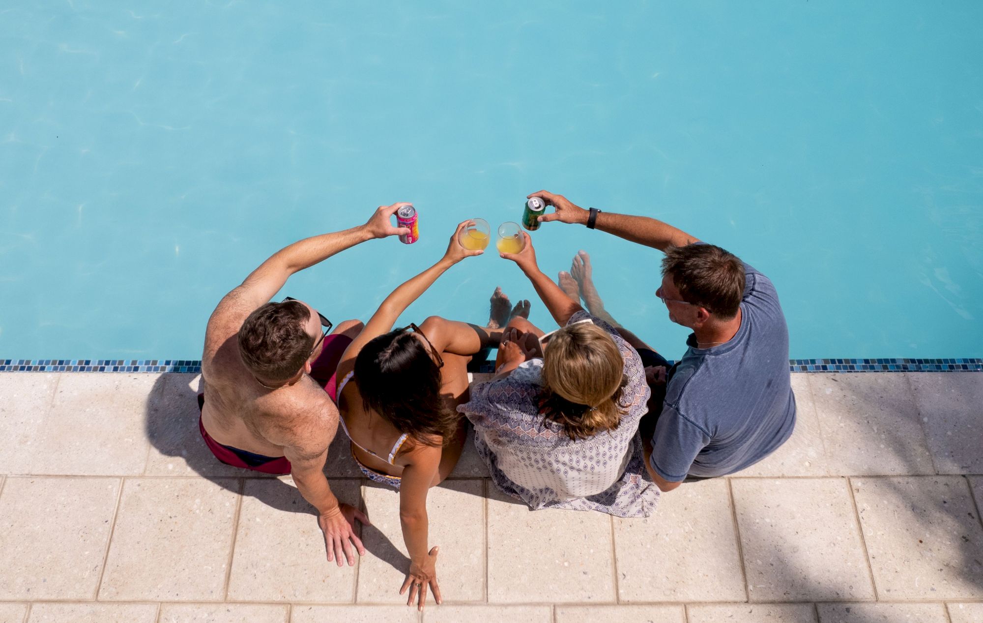 Four people are sitting by a pool, holding drinks and toasting together, with their legs dipped in the water.