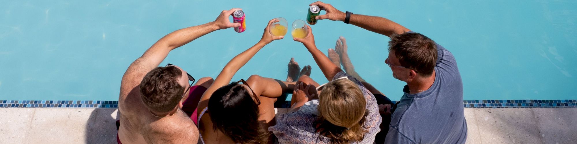 Four people are sitting by a pool, holding drinks and toasting together, with their legs dipped in the water.