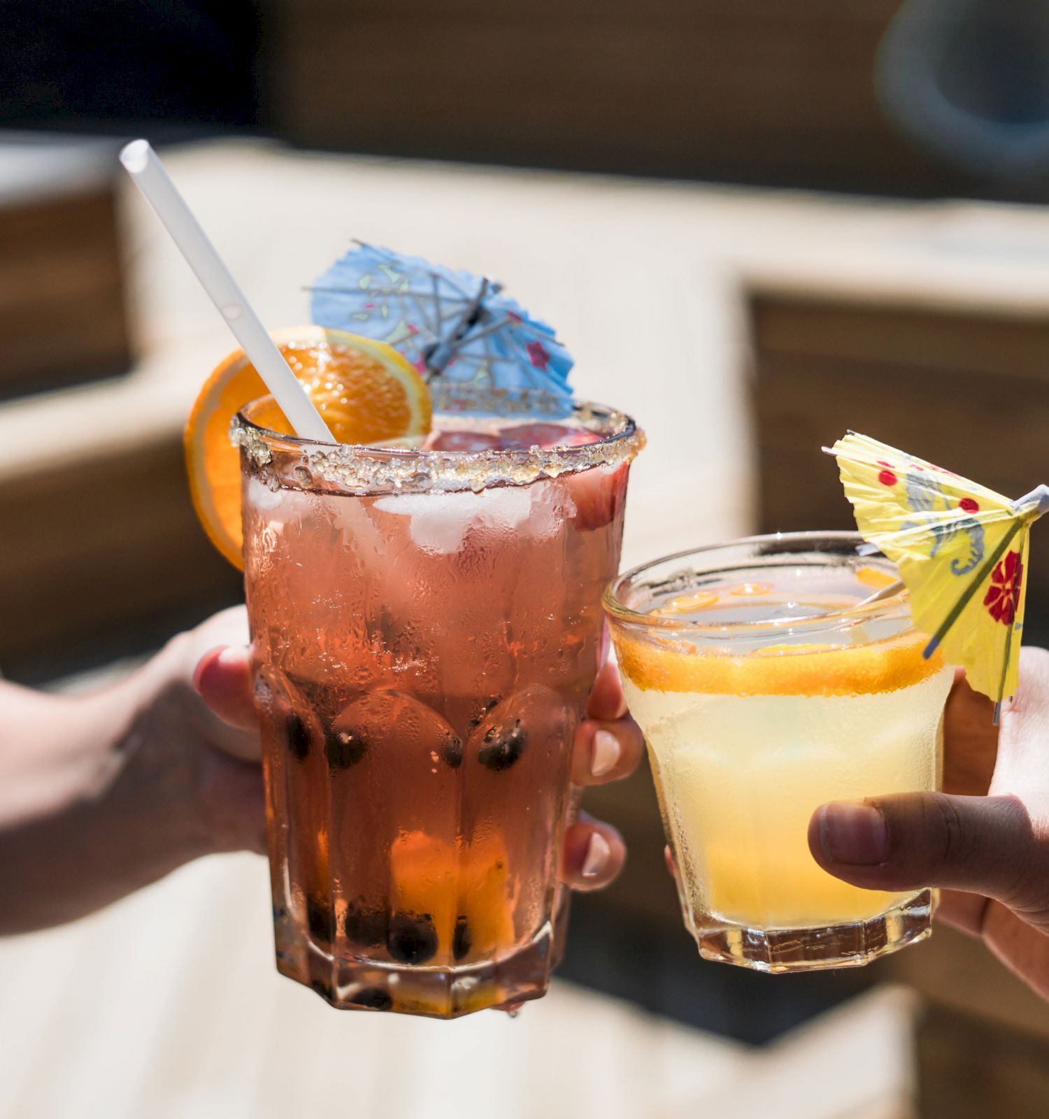 Two people clinking glasses with colorful cocktails, each adorned with a small decorative umbrella and a slice of citrus fruit.