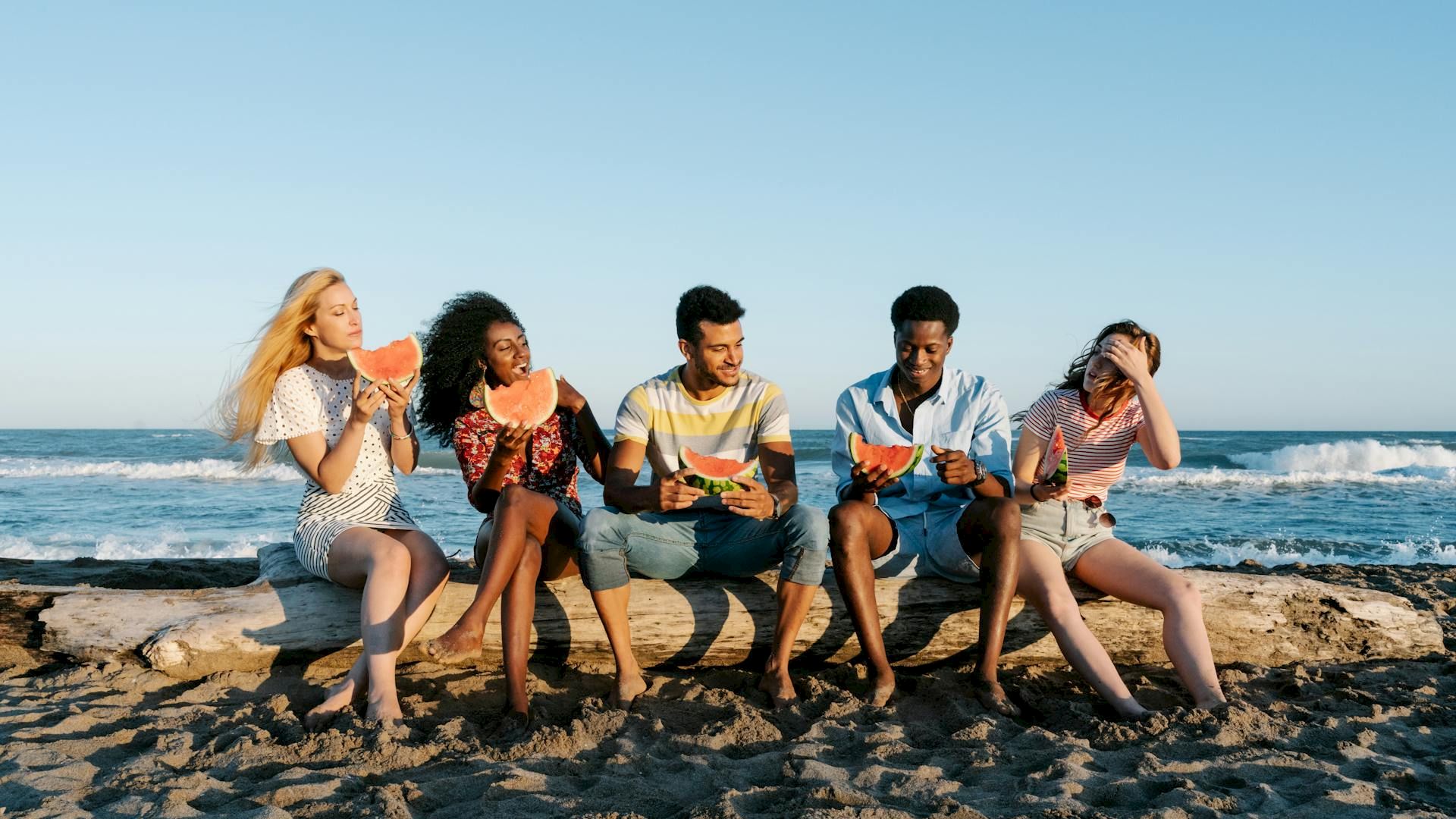 Five people sitting on a beach log, eating watermelon, with the ocean in the background.