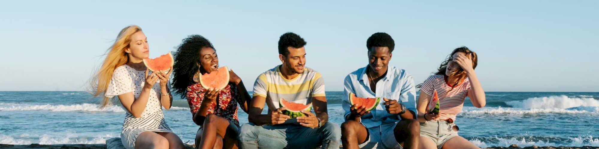 Five people sitting on a beach log, eating watermelon, with the ocean in the background.