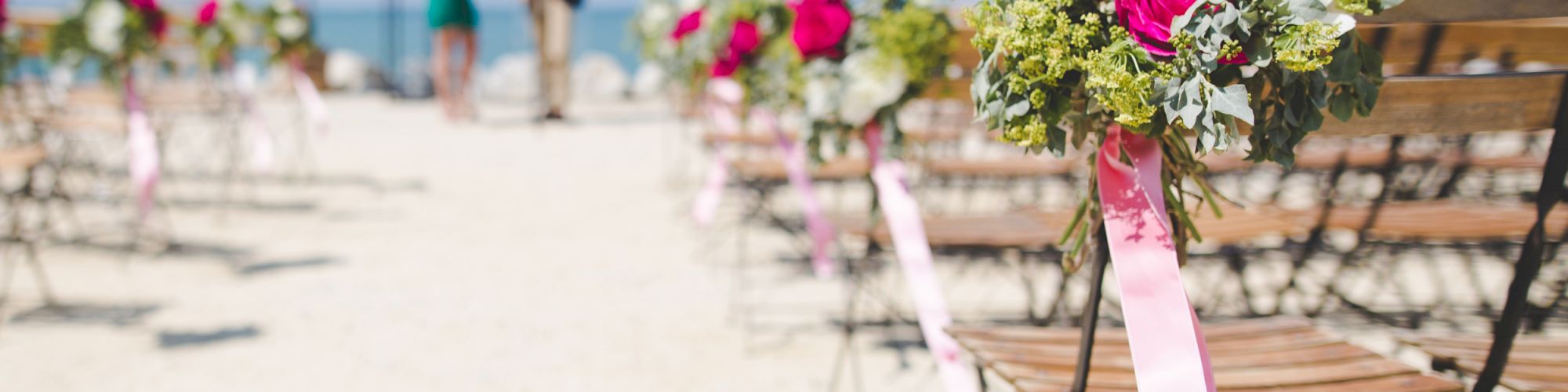 Outdoor wedding setup with rows of chairs decorated with flowers and pink ribbons, overlooking a beach with people blurred in the background.