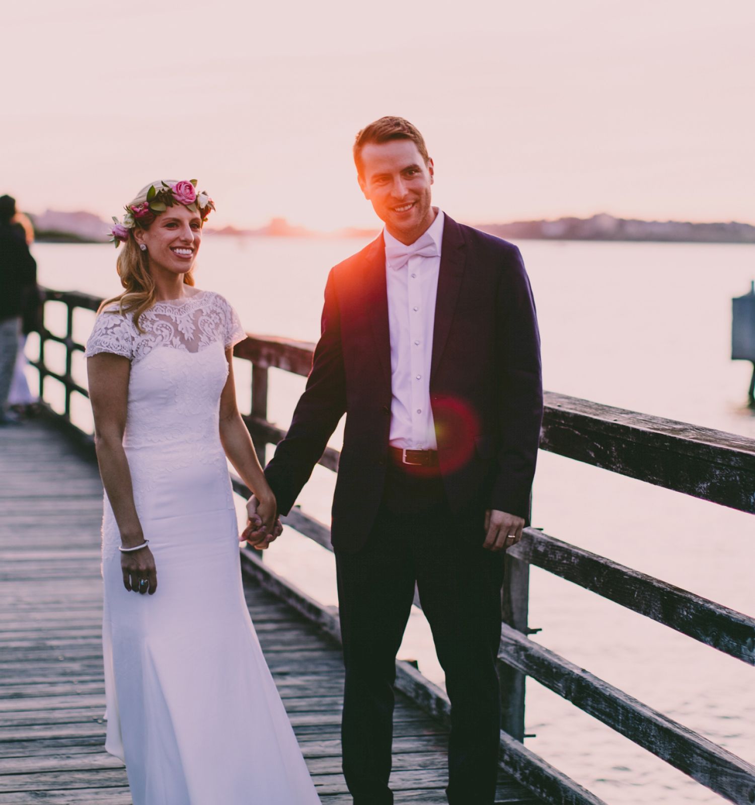 A couple in wedding attire holds hands on a wooden pier at sunset, with water and some structures in the background.