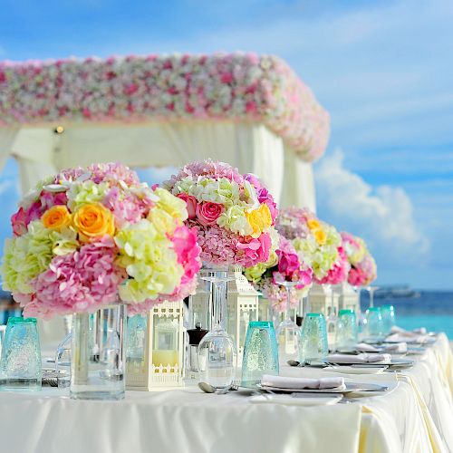 An elegantly decorated outdoor table with colorful floral arrangements and ocean views, set under a canopy adorned with flowers, ready for a special event.