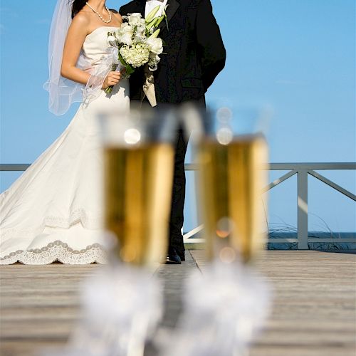 A bride and groom stand on a deck, smiling at each other, with two champagne flutes in the foreground.