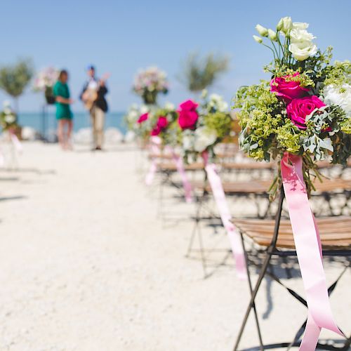 Outdoor wedding setup with wooden chairs and floral decorations on a sandy surface, with a couple in the background.