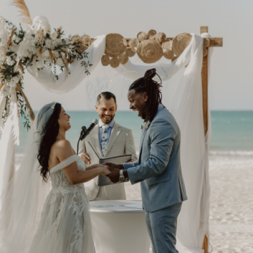 A couple is exchanging vows at a beach wedding, with an officiant standing behind them under a decorated arch, all smiling warmly.