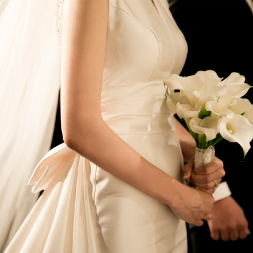 A bride in a white dress holds a bouquet of white flowers, with a veil draped over her shoulder, standing next to a groom in a black suit.