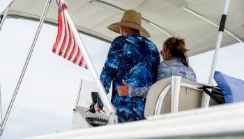 A man and woman are on a boat, with the man steering. They are wearing sun-protective clothing. An American flag is visible on the boat.