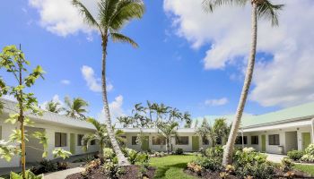 A courtyard with lush greenery, palm trees, and single-story white buildings under a bright blue sky with scattered clouds.