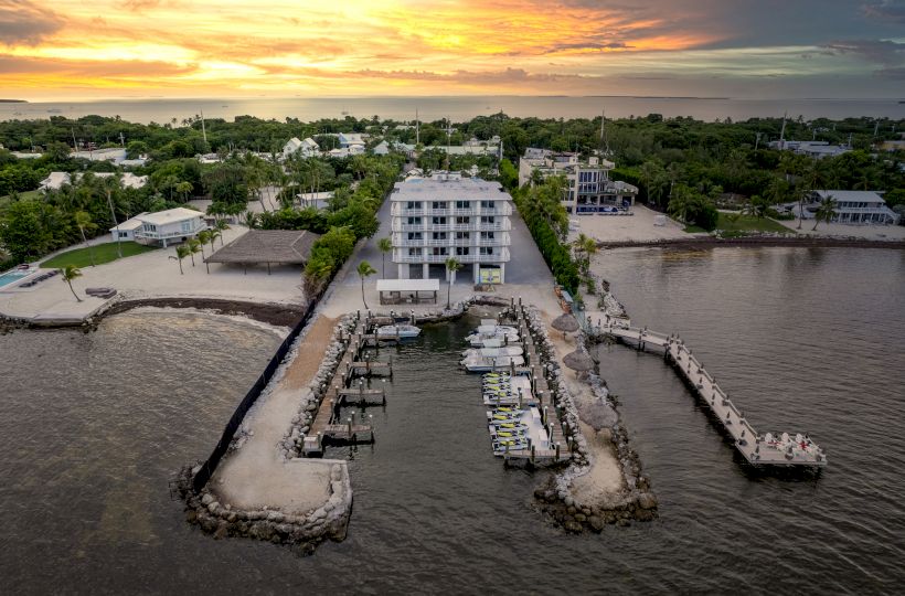 An aerial view of a waterfront property with a centrally located building, boat docks, surrounding greenery, and a stunning sunset in the background.